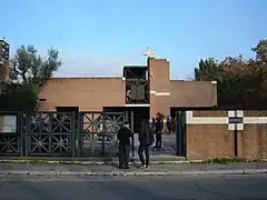 Une église à l'architecture moderne, en brique. Elle est surmontée d'une croix carrée et accompagnée d'un portail en fer forgé qui donne sur la rue.