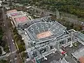 Vue plongeante du court Suzanne-Lenglen. Les tribunes sont vides et grises, la surface du terrain semble très claire. Sur la partie supérieure, on distingue l'A13 avec une circulation fluide. De chaque côté du court, des échaffaudages sont en cours d'assemblage ou de désassemblage. Plusieurs courts plus modestes sont visibles derrière le Suzanne-Lenglen. Le boulevard d'Auteuil, bordé d'arbres, longe les courts.