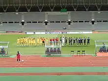Deux équipes de football, jaune à gauche, bleu marine à droite, alignées sur un terrain de sport, devant une grande tribune.