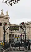 Photographie d'une bouche de métro occupant le bas de l'image, tandis que le haut de l'image montre deux allégories féminines de villes allemandes, Berlin et Cologne, surmontées de feuilles de platane se détachant sur fond de ciel blanc.