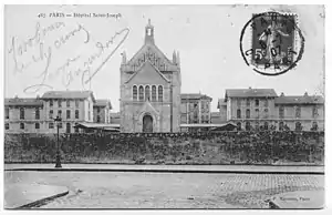 Carte postale en noir et blanc d'un groupe de bâtiments séparé d'une rue pavée par un mur, avec cachet postal portant la date du 8 juillet 1907.