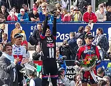 Photographie en couleur représentant, au premier plan, devant la foule, au centre le vainqueur levant le trophée, un pavé, au dessus de sa tête, encadré par les deuxième et troisième portant un bouquet de fleur