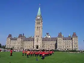 Photo en couleurs de militaires en uniformes rouges sur une place devant le bâtiment du Parlement du Canada