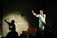 Photographie couleur d'un homme récitant dans un micro, et éclairé par un projecteur qui projette son ombre sur un mur latéral.