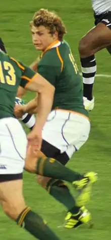 Le joueur, de profil, est en pleine action avec le maillot vert et or de l'équipe d'Afrique du Sud.