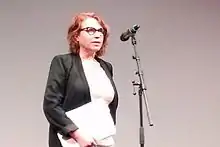 Plan américain d'une femme de 55 ans, rousse, portant des lunettes, devant un micro.