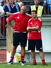 Deux hommes observent un match de rugby depuis le bord du terrain.