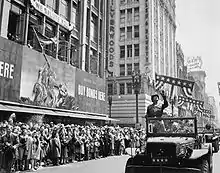 Patton en uniforme se tient debout dans un véhicule militaire. Des spectateurs saluent son passage tandis que des cotillons sont jetés depuis les gratte-ciels bordant la rue.