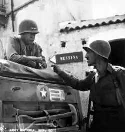 Patton portant un casque avec trois étoiles sur le devant et un uniforme d'officier supérieur discute depuis un véhicule avec un homme casqué en tenue de combat moins formelle. À l'arrière-plan, un panneau accroché sur un mur donne la direction de Messine.
