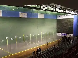 Vue de l'intérieur d'une salle de sport, à gauche un terrain de pelote basque, à droite des tribunes.