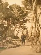 Un homme en costume sombre et chapeau dans un parc avec de grands pins.