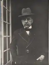 Homme avec chapeau et veste, nœud papillon devant une porte vitrée ouverte.