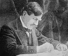 Paul Bourget vers 1893, assis à une table en train d'écrire, porte-plume dans la main droite et bras gauche appuyé sur le bureau, visage penché et regard concentré sur son manuscrit.