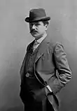 Paul Bourget, jeune homme, chapeau, cravate à carreaux, costume et gilet clair, est photographié mains dans les poches. Sur le côté gauche de son gilet, une montre est reliée à une poche par une chaine.