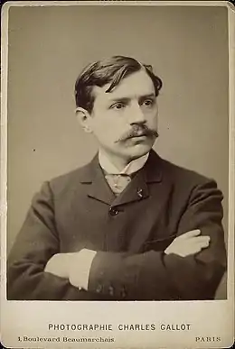 Photographie en noir et blanc d'un homme moustachu, les bras croisés.