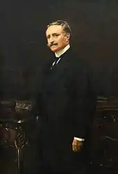 Portrait en couleur et de trois-quarts d’un homme debout, moustache et cheveux châtains grisonnants, portant une chemise blanche à faux col, une cravate, un veston et une veste de couleur noire