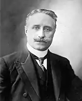 Photo en noir et blanc et de face d'un homme à la moustache et aux cheveux grisonnants, portant une chemise à faux col, une cravate, un veston et une veste épaisse