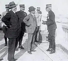 Photo d'un homme en complet gris à rayures, portant un chapeau mou et des gants, et échangeant avec un général de brigade