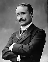 Photo en noir et blanc d’un homme aux bras croisés, avec moustache en croc et cheveux châtains, portant une cravate, une veste et une chemise blanche au col relevé