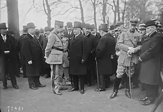 Photo en noir et blanc d'un groupe de personnes avec, au centre, un homme à la moustache et aux cheveux blancs, portant un haut-de-forme et conversant avec un militaire portant les galons de général de division