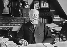Photo en noir et blanc d'un homme dégarni et à la barbe blanche, assis à un bureau, vêtu d'une redingote, d'une chemise blanche et d'une cravate