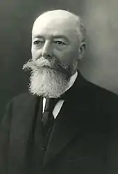 Portrait photographique d'un homme aux cheveux et à la barbe grisonnants, vêtu d’une chemise blanche, d’un veston, d'une veste et d'une cravate foncés