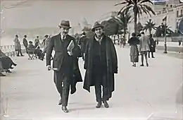 Photo noir et blanc de deux hommes marchant, l'un moustachu en costume, l'autre en long manteau et chapeau, l'air fatigué