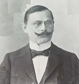 Portrait en noir et blanc d'un homme aux cheveux bruns et courts, habillé d'un costume sombre, arborant un pince-nez, une moustache en guidon et un nœud-papillon.