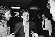 Paul McCartney jeune, photo en noir et blanc, en train de se faire interroger par des journalistes dans le couloir d'un immeuble