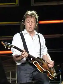 Paul à la guitare, debout en chemise blanche et bretelles rouges, jambes légèrement écartées
