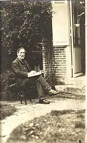 Photographie de Paul Milliet, plus âgé que durant les évènements de la Commune. Il est assis sur une chaise, dehors devant une maison, et tient un livre entre ses mains.
