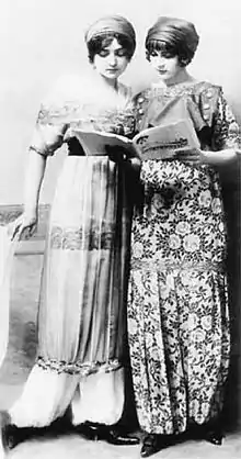Photographie en noir et blanc de deux femmes feuilletant un magazine, l'une en robe et l'autre en pantalon bouffant, portant toutes les deux des chapeaux.