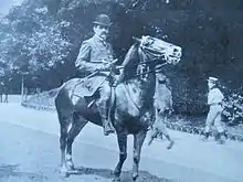 Homme à cheval dans une allée de parc public avec passants. Le cavalier porte un chapeau.
