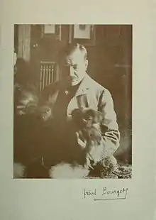 Un homme moustachu est assis avec trois peits chiens qu'il regarde. La photographie est signée de la main de Paul Bourget.
