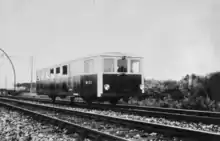 Photographie en noir et blanc d'un autorail bicolore (haut de caisse blanc et bas de caisse foncé) de forme parallélépipédique sous une caténaire dont les poteaux ont la forme d'une ogive, caractéristiques du Midi.