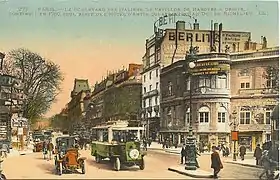 Carte postale colorisée représentant une avenue bordée d'immeubles, avec des passants et des automobiles