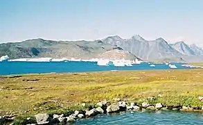 Photographie en couleurs d'un terrain herbacée entouré de plans d'eau, un massif montagneux visible en arrière-plan.