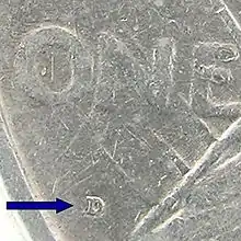 Gros plan sur une pièce de monnaie : l'inscription ONE est visible ainsi que la lettre D majuscule vers laquelle pointe une flèche.