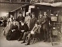 Photographie en noir et blanc d'un groupe posant sur un navire, trois personnages sont assis : une dame aux cheveux blancs, un homme à la longue barbe blanche et un jeune homme portant un chapeau et regardant l'horizon.