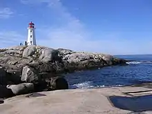 Photo couleur. La mer baigne au premier plan une plage de sable gris puis une avancée de rochers sur laquelle se trouve un phare.