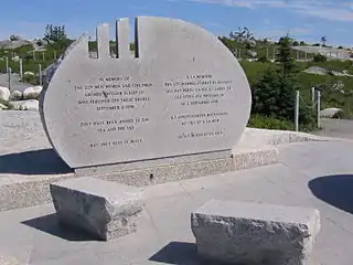 Photo couleur d'une pierre commémorative grise de forme demi-circulaire. Des petits bancs de pierre sont présents devant le mémorial, qui comporte des inscriptions en français et en anglais.