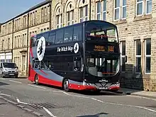 La photographie montre un bus à deux étages stationné dans une rue devant un immeuble en pierre blanche à trois étages. Il est peint en noir, avec deux bandes grise et rouge. Sur le côté est dessiné une sorcière volant sur son balai devant la lune.