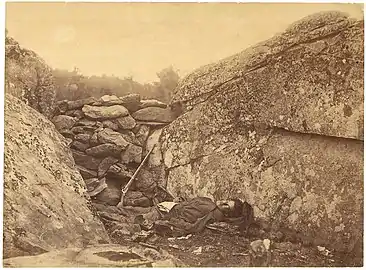 Photographie en noir et blanc d'un soldat mort, allongé sur le dos, légèrement tourné vers l'objectif, au milieu de rochers