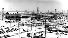 Vue étendue d'un chantier de construction. Aux alentours, on peut voir de nombreuses voitures garées, des échafaudages et des grues.