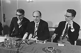 Andréas Papandréou flanqué de deux hommes assis à une table devant des microphones