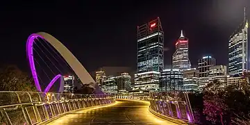 Le passage du pont, éclairée de lumières rasantes, de nuit. Sur le coté une arche violette jaillit dans le ciel. Au bout de la perspective des buildings qui se donnent des allures de gratte-ciel.