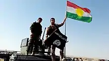 Photographie couleur. Deux hommes en treillis se tiennent face caméra, debout sur un véhicule militaire. L'homme à gauche arbore un écusson kurde sur la manche droite de sa veste. Ce même homme désigne de sa main gauche un drapeau du Kurdistan hissé sur le véhicule tandis qu'il tient dans sa main droite le drapeau en berne de l'état islamique. Ciel dégagé en arrière plan.