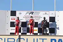 Photographie de trois hommes sur un podium, debouts. Les deux de gauche sont en rouge, celui de droite en noir.