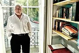 Photographie couleur. Homme d'âge mur, debout devant une fenêtre à côté d'une bibliothèque.