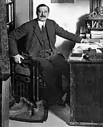 photographie en noir et blanc. Un homme prend la pose assis à son bureau, une main sur l'accoudoir, une main sur le plateau.